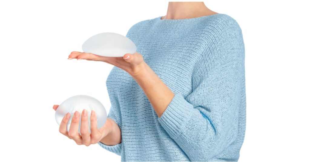 A person in a light blue sweater holds two breast implants, one in each hand, against a white background. (model)